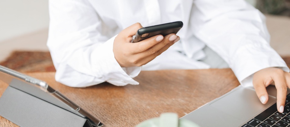 A person working remotely at a desk using a smartphone and laptop.