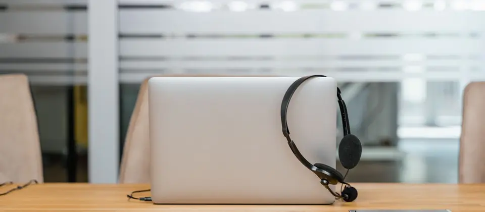 A closed laptop resting on a wooden desk with a professional headset draped over it in a modern office setting, representing remote virtual assistant and support equipment